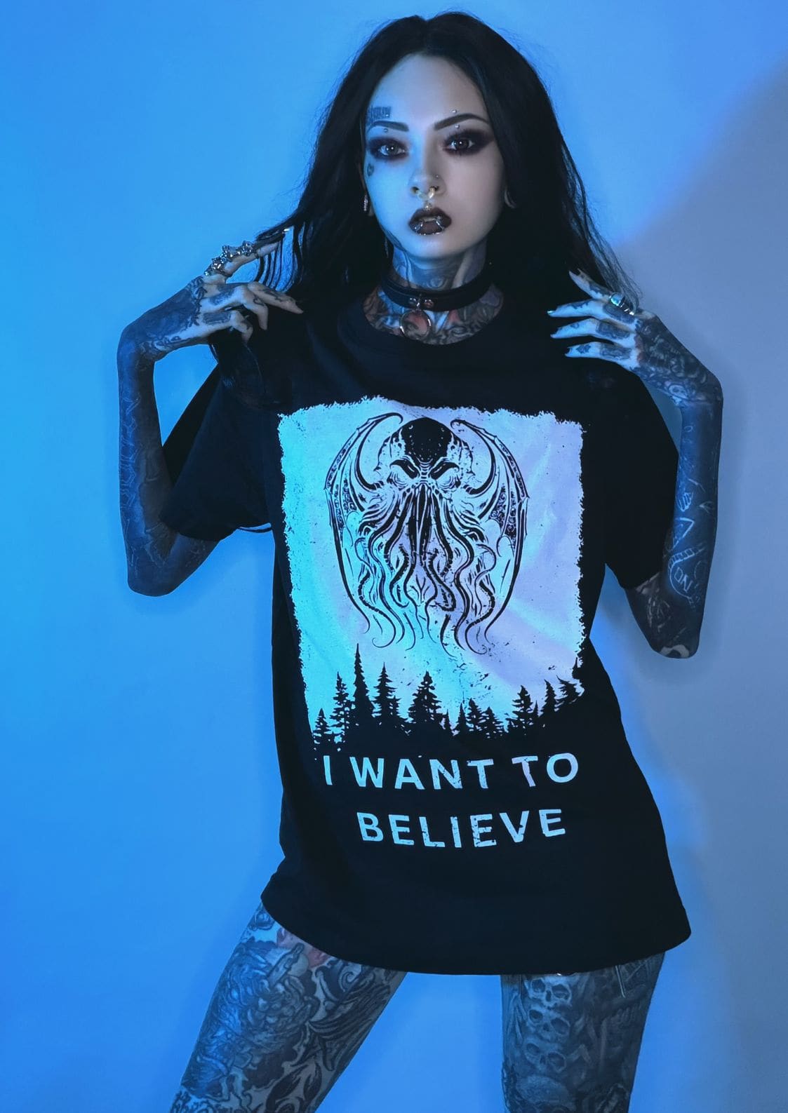 Schwarzes T-Shirt mit einem weißen Grafikmotiv eines octopus-ähnlichen Wesens mit Flügeln über einer Silhouette eines Waldes und dem Text ‚i want to believe‘ in fetten, blockartigen Buchstaben.