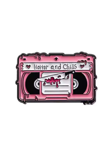 Pink enamel pin in cassette tape shape with horror and chills label and knife through blood red film for gothic dark fashion