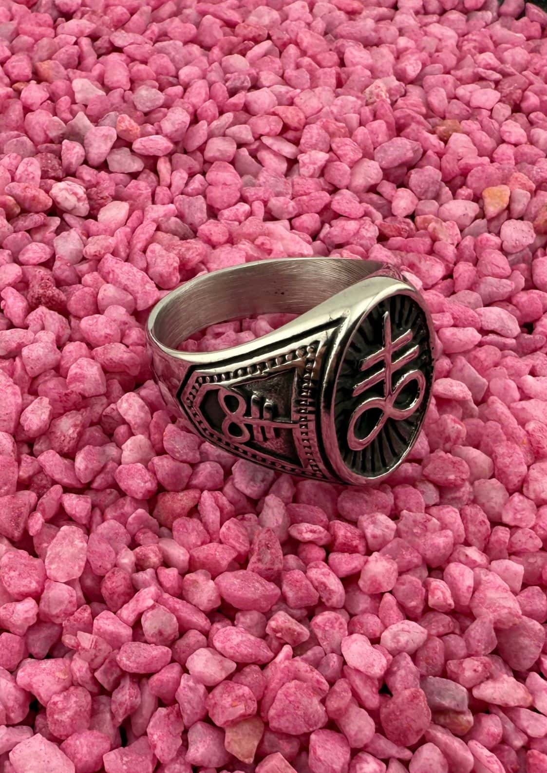 Silberner Ring mit schwarzem Design und Text, auf rosa Kies gesetzt.