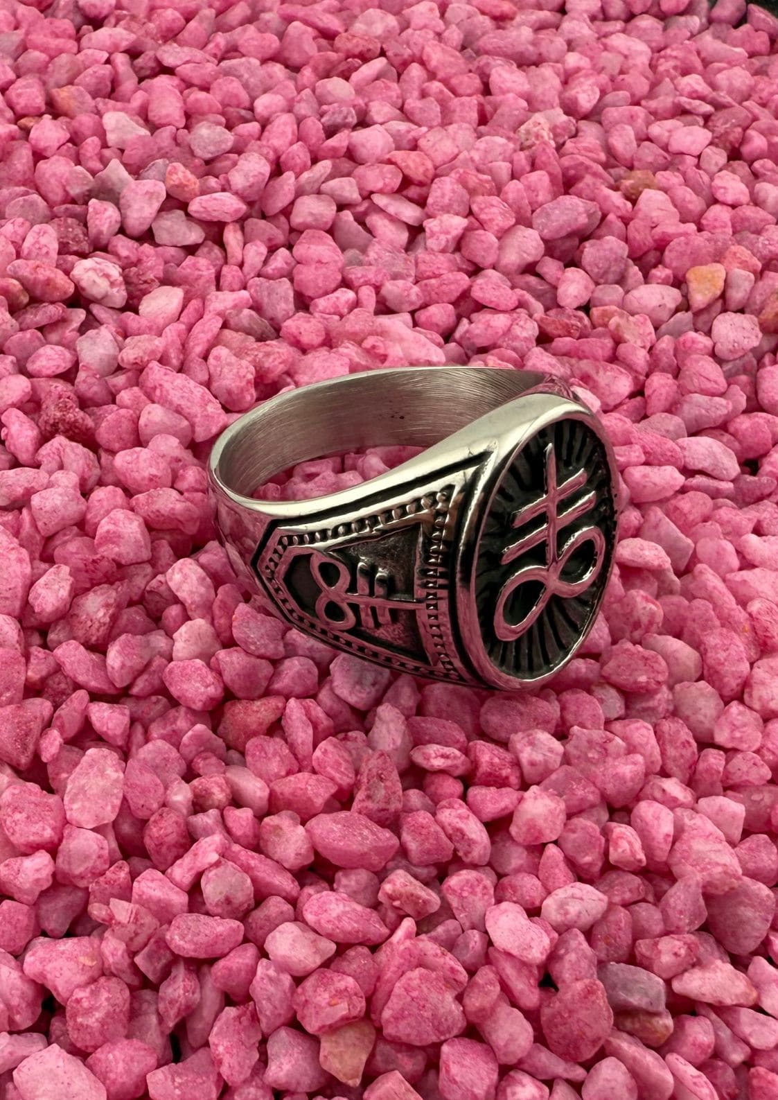 Silberner Ring mit schwarzem Design und Text, auf rosa Kies gesetzt.