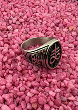 Silberner Ring mit schwarzem Design und Text, auf rosa Kies gesetzt.