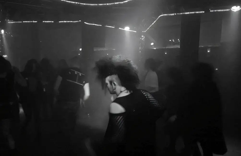 Eine Person mit einem Irokesenschnitt und dunkler, strukturierter Kleidung steht in der Mitte eines spärlich beleuchteten Clubs.