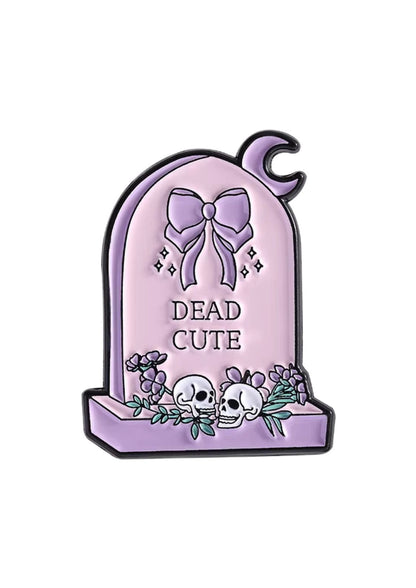 Ein Lavendel-Farbenes Email-Nadel in Form eines Grabsteins mit einer Schleife, Totenköpfen, Blumen und den eingravierten Worten „dead cute“.