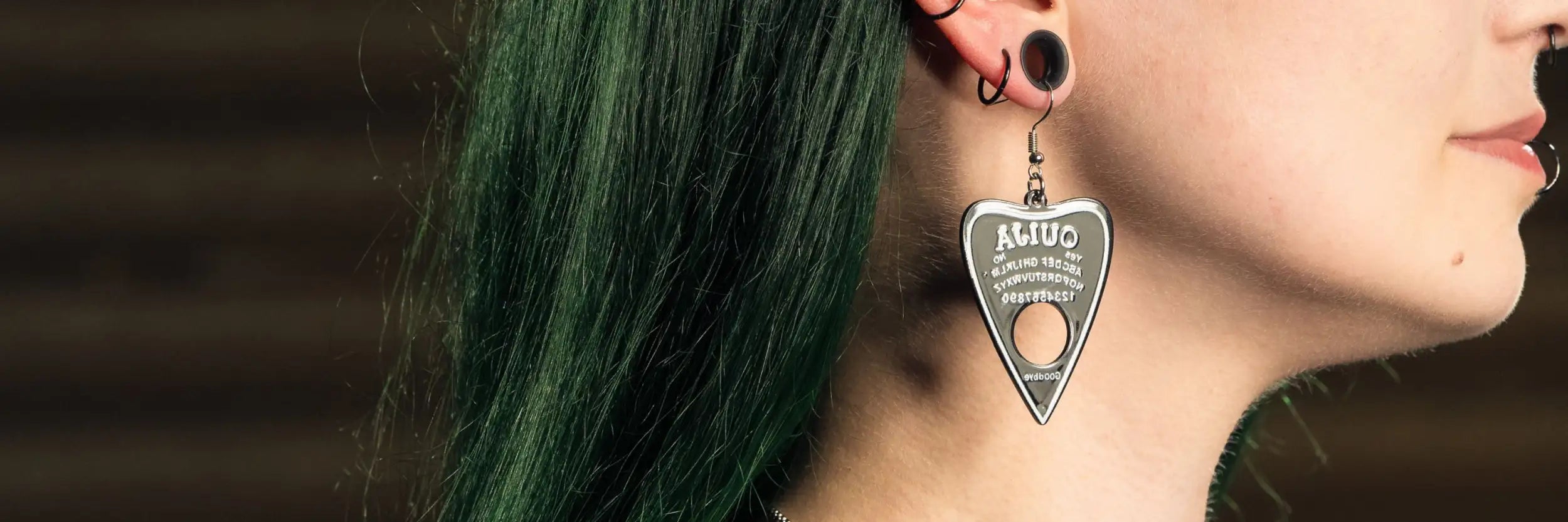 Silbernes metallisches dreieckiges Anhänger-Ohrgehänge mit graviertem Text und einer zentralen runden Öffnung, gestaltet, um einem Ouija-Brett zu ähneln.
