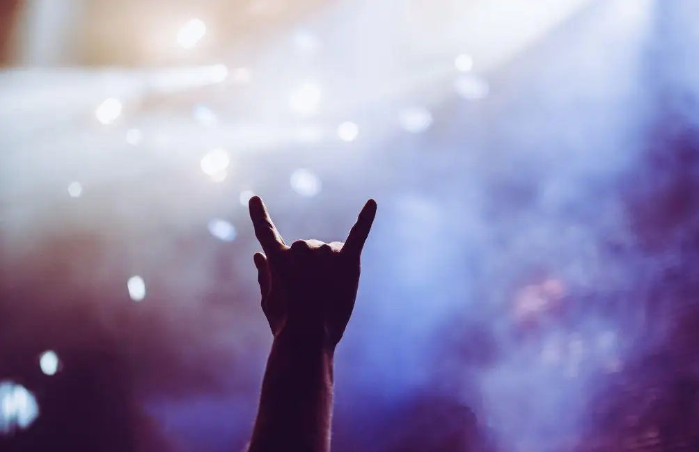 Eine silhouettierte Hand, die eine Rock-Geste gegen einen Hintergrund aus lebendigen Bühnenlichtern und Nebel macht.