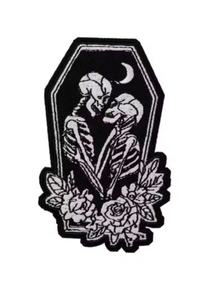 Schwarz-weiß gestickter Aufnäher mit zwei sich umarmenden Skeletten in einem Sarg mit Rosen.