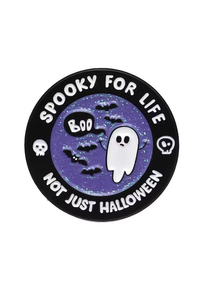 Runde schwarze Emaille-Anstecknadel mit lilafarbenem Glitzerhintergrund, auf dem ein Geist, Fledermäuse und Totenköpfe abgebildet sind.