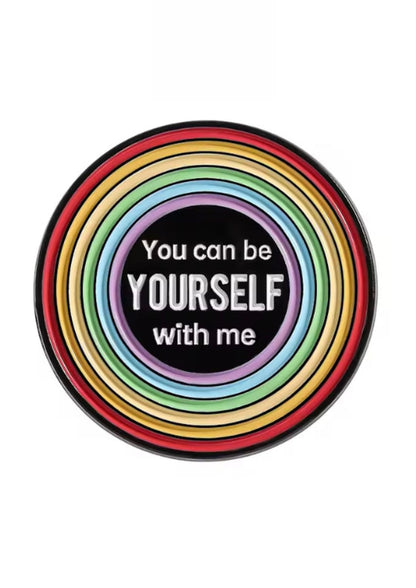 Regenbogenfarbene runde Anstecknadel mit dem Text ’Du kannst mit mir du selbst sein’ auf schwarzem Hintergrund.